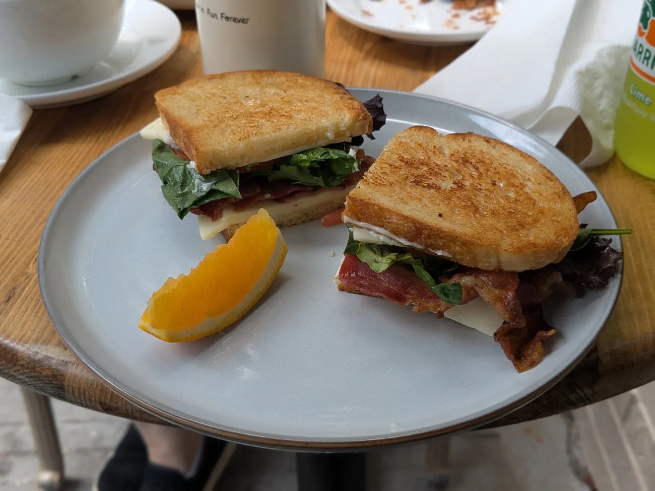 Toasted sandwich cut in half with bacon, cheese and greens, served on a plate with an orange wedge